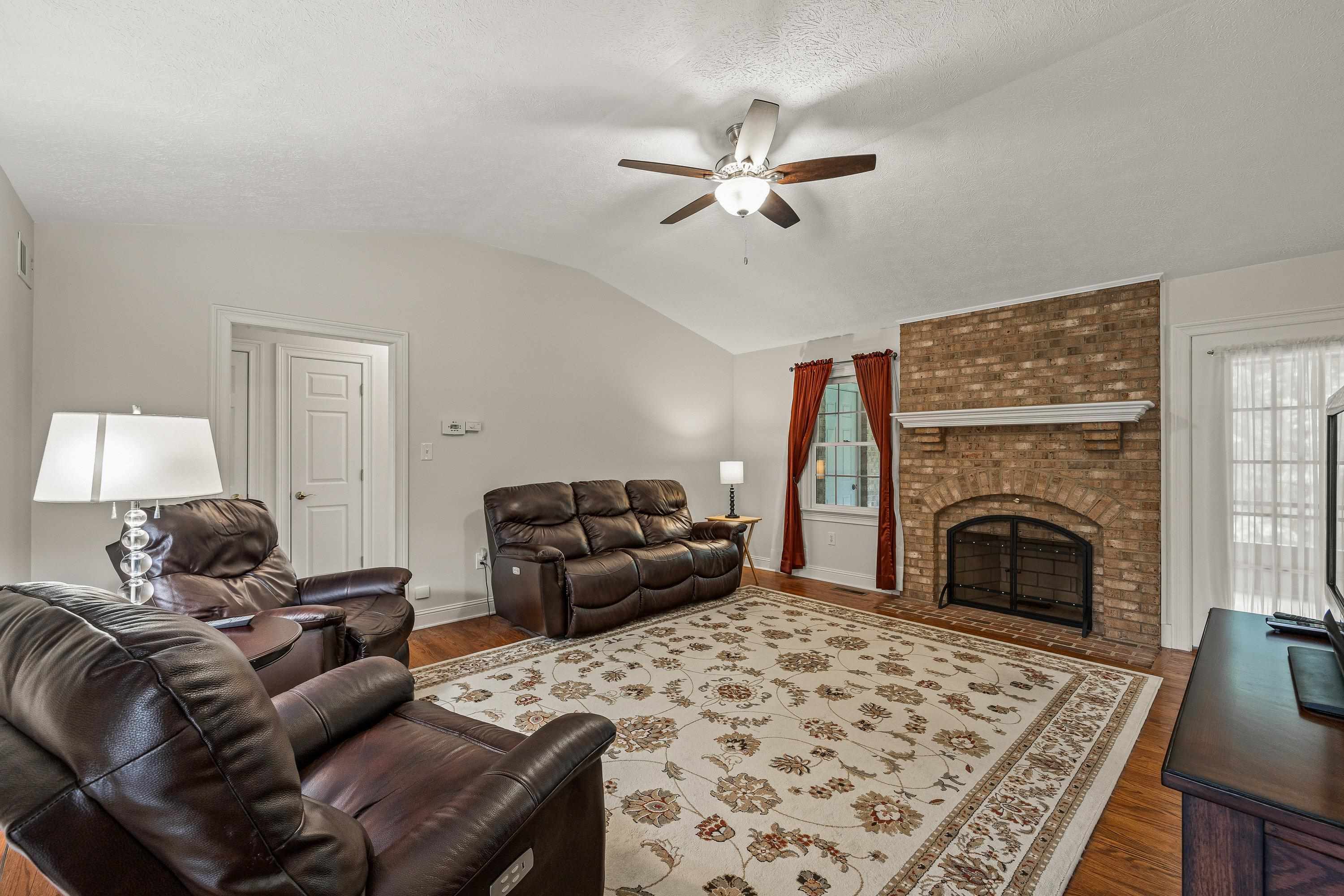 Open and bright, this living room space contains a brick wood fireplace, hardwood flooring, large windows, ceiling fan, and an abundance of space for gathering with family and friends!
