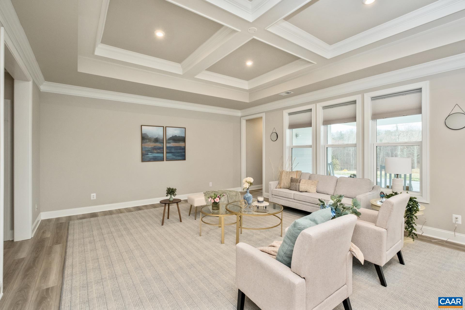 The living room features a raised tray and coffered ceiling, triple windows to allow in plenty of sunlight, and a custom-made rug that conveys.  In view to the far right is the private hallway leading to the primary suite.