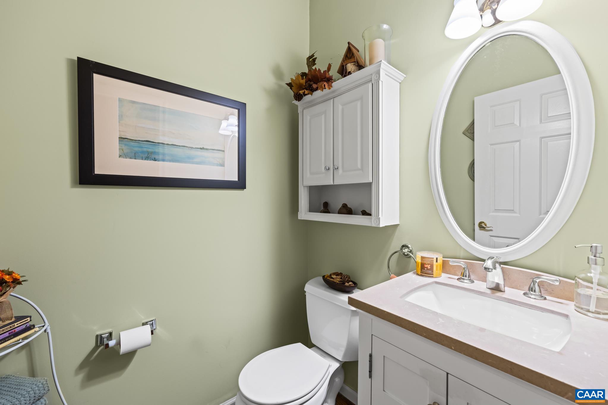 Bright powder room on the 1st floor features a classic white vanity with a light countertop and oval mirror, complemented by soft, pale green walls. The wall-mounted cabinet offers storage, and the space is accented with a framed coastal landscape print. Bright powder room on the 1st floor features a classic white vanity with a light countertop and oval mirror, complemented by soft, pale green walls. The wall-mounted cabinet offers storage, and the space is accented with a framed coastal landscape print.