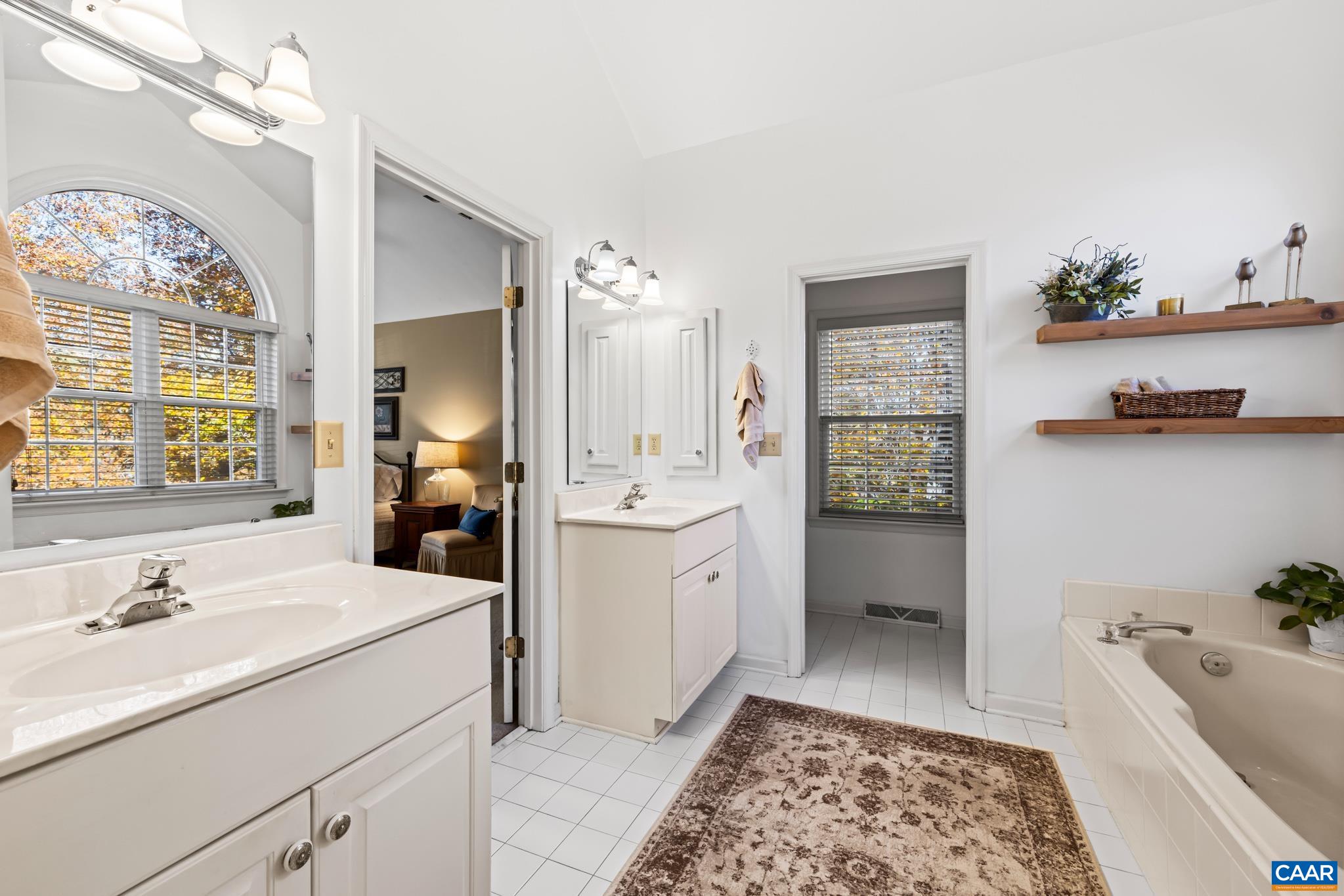 Relax in this bright primary bath with dual vanities, white tile flooring, a soaking jetted tub, and a large arched window overlooking vibrant foliage. Relax in this bright primary bath with dual vanities, white tile flooring, a soaking jetted tub, and a large arched window overlooking vibrant foliage.