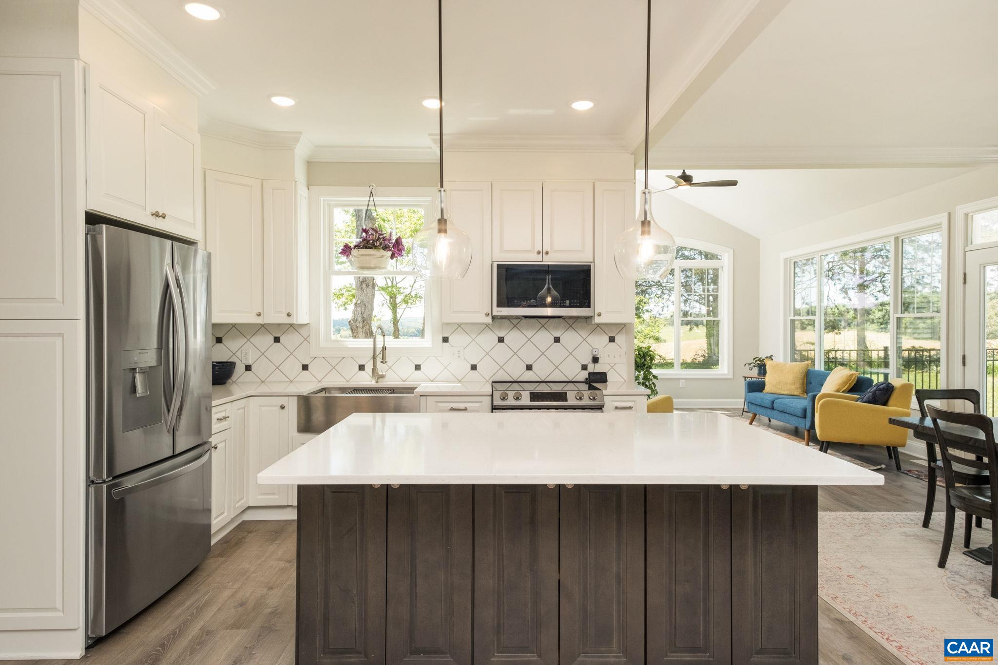 The kitchen features a large quartz countertop island with plenty of cabinetry, coffered ceiling, tile backsplash, and cabinetry panels around the refrigerator.  Out of view is the walk-in pantry and welcome center, to the left of the kitchen.