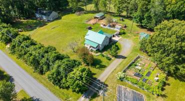 Aerial view showcasing the established homestead setting with home, outbuildings, garden areas, and open green space surrounded by countryside.