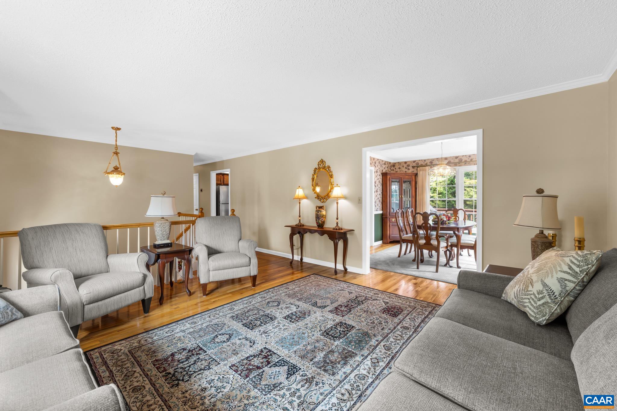 Bright, welcoming living room featuring gleaming hardwood floors, classic crown molding, and warm accent lighting. Wide cased openings create seamless flow into the adjacent formal dining area—perfect for effortless hosting!