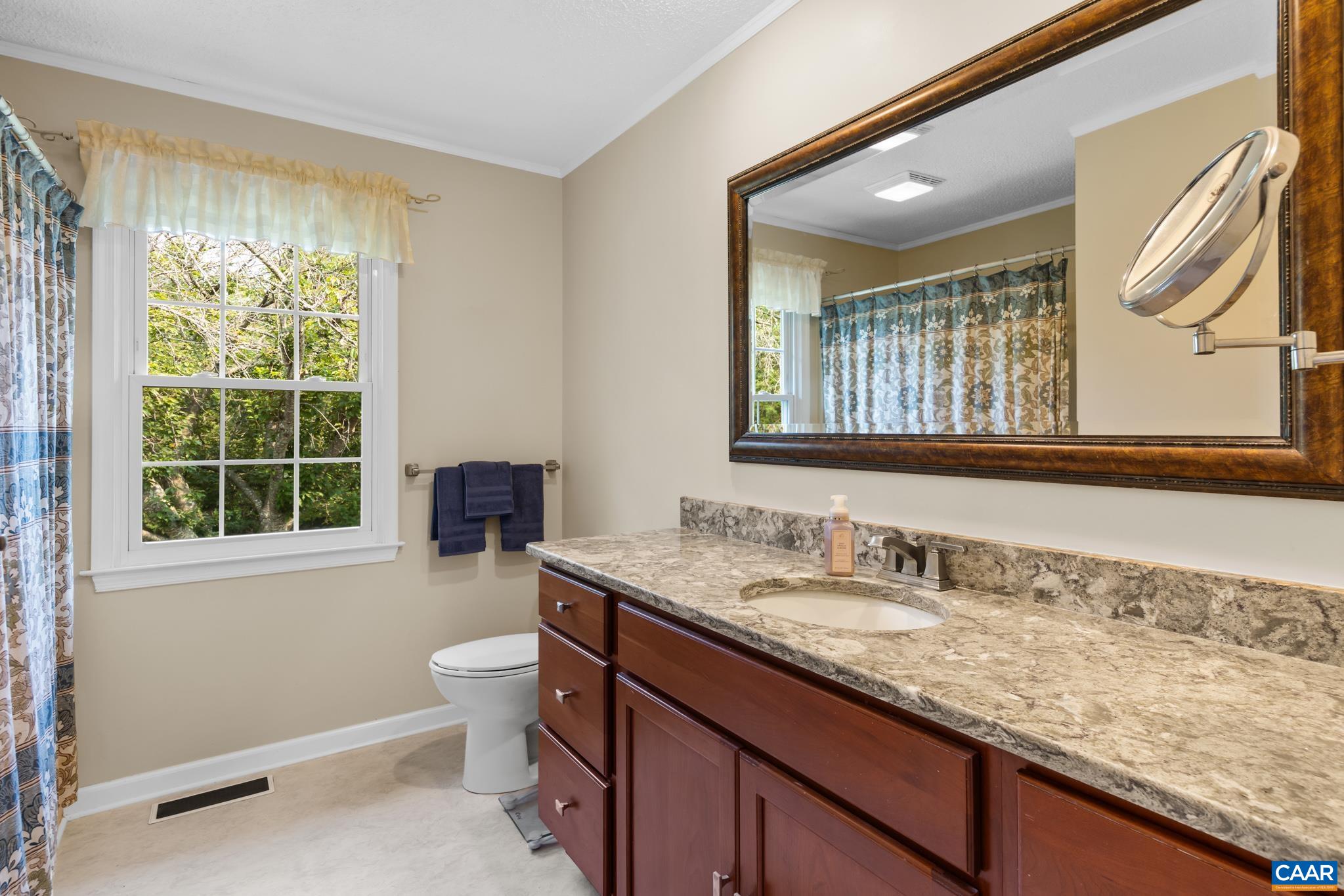 This bright and spacious bath features a rich wood vanity with a beautiful granite countertop, a large framed wall mirror, a mounted magnifying mirror, and a sunlit window. Clean, stylish, and functional—ready for your daily routine!