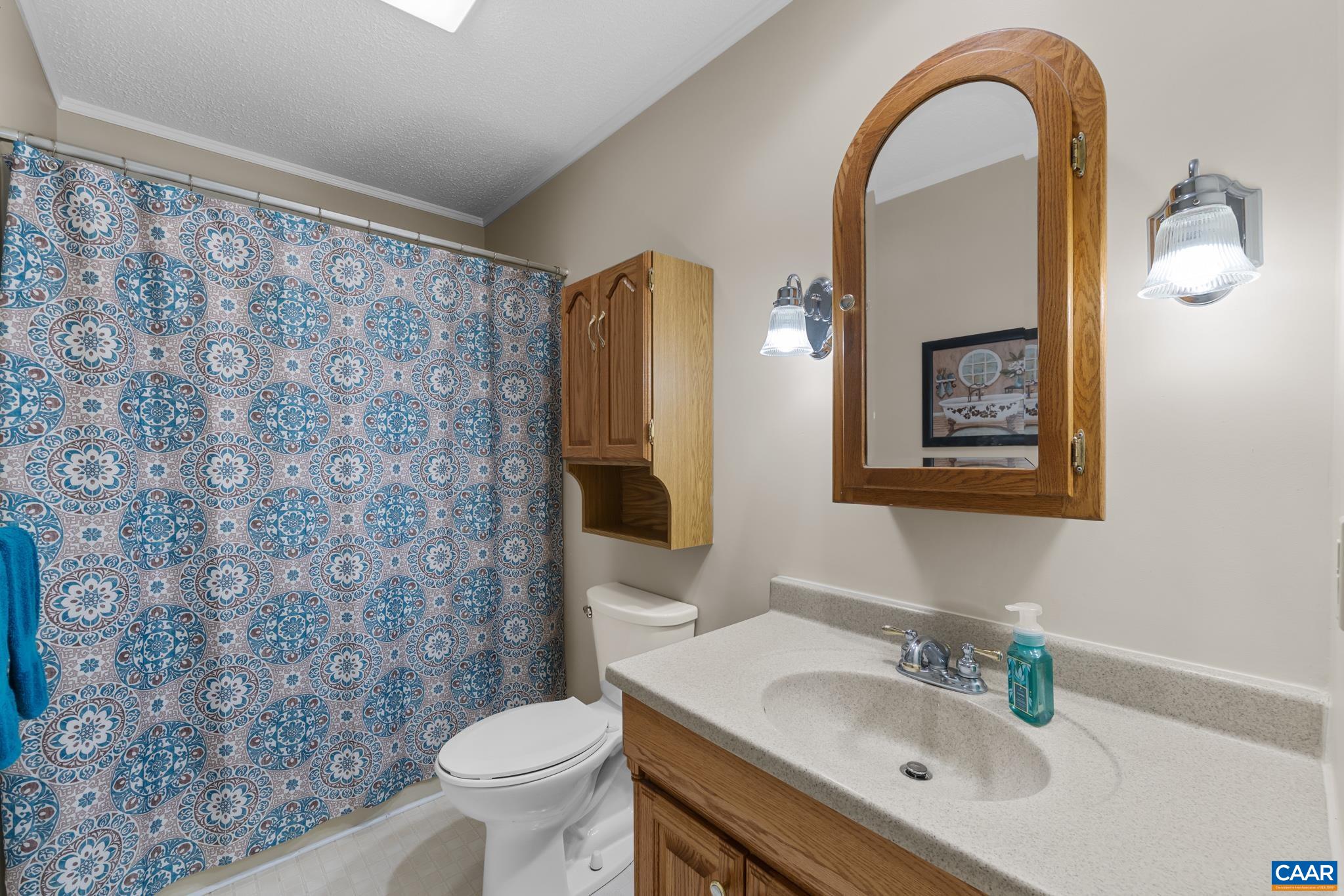 A classic and functional full bathroom! Features a warm oak-finished vanity, an arched wooden medicine cabinet mirror, convenient over-the-toilet storage cabinet, and classic sconce lighting. A clean, inviting space perfect for everyday ease!