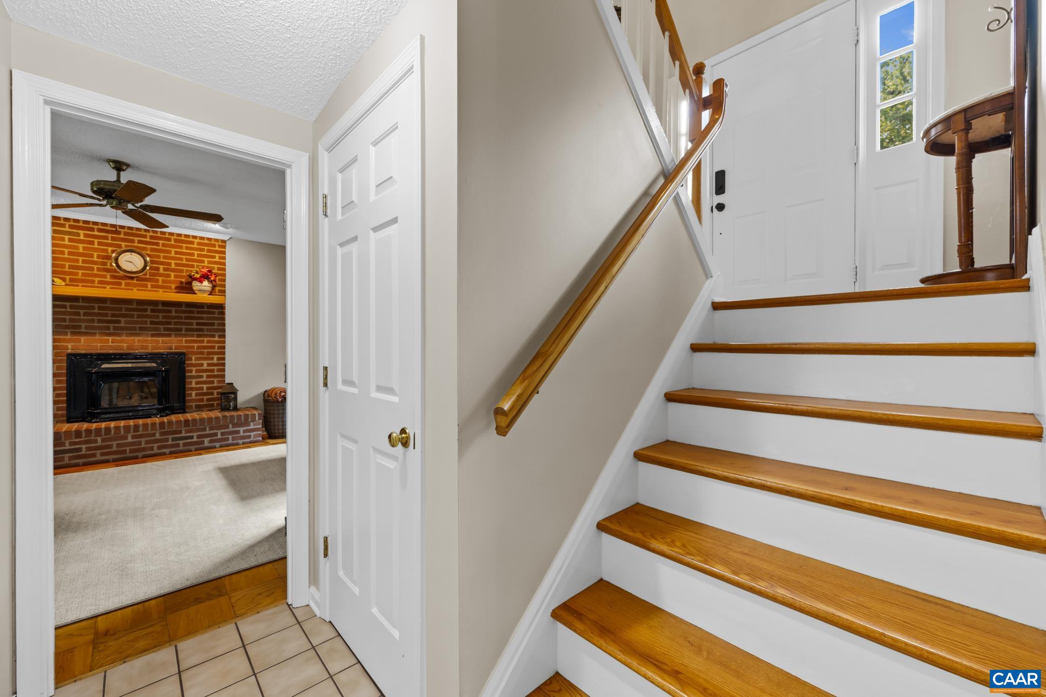 An inviting split-foyer entryway! Features warm hardwood stairs with crisp white risers, clean white trim, and a bright view looking toward the front door on one side and a cozy brick fireplace den on the other. Seamless flow throughout!