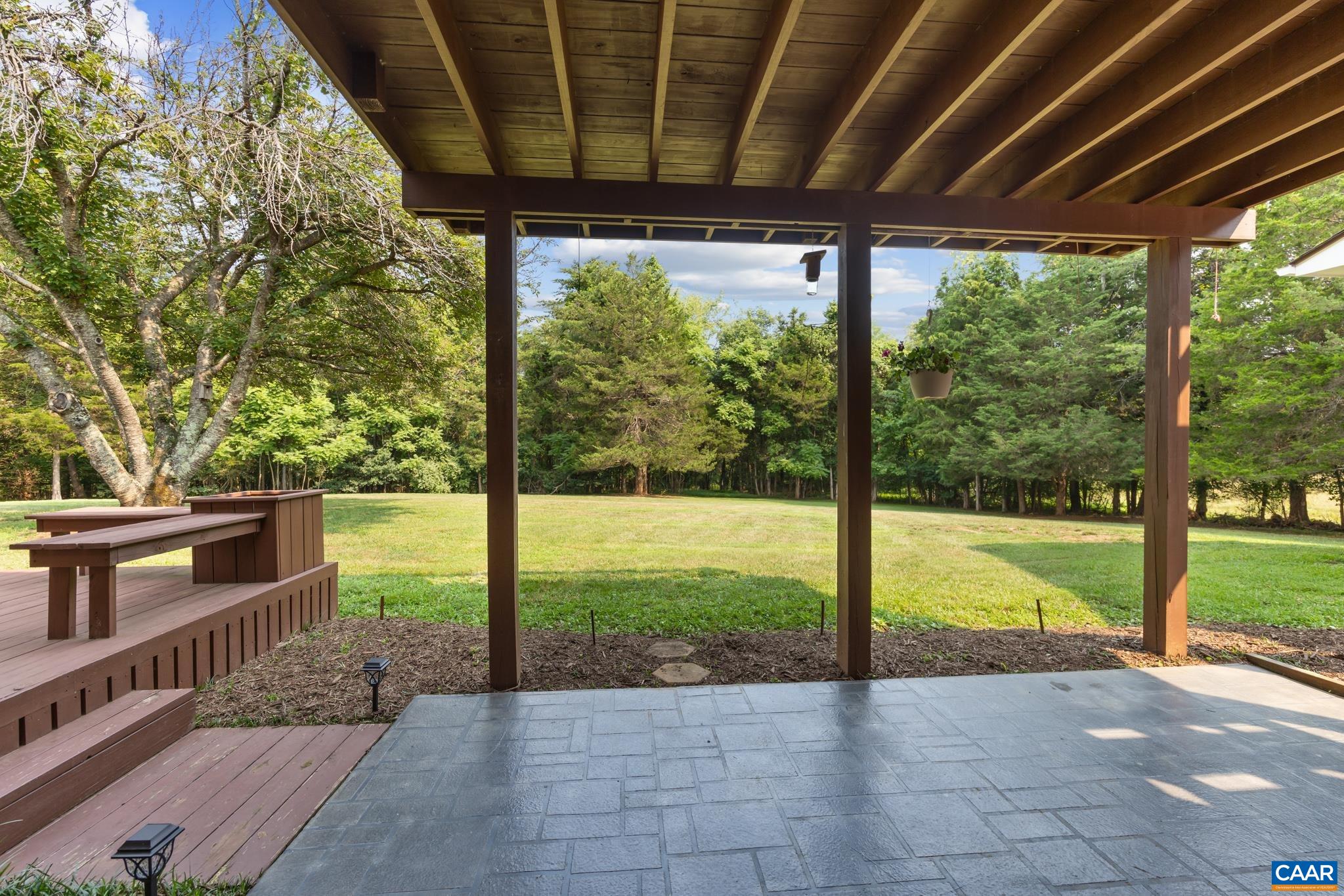 Enjoy sheltered outdoor living under this covered patio! Features stamped concrete flooring, wooden support posts, and steps leading to an adjacent deck, all overlooking a vast, lush lawn framed by mature trees. Perfect for shade and relaxation!