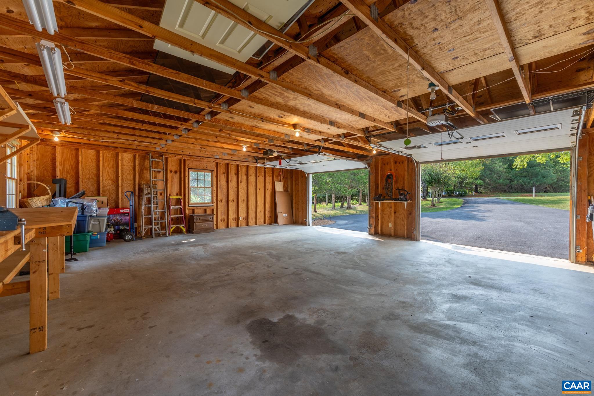 Spacious multi-bay garage interior! Features solid concrete flooring, exposed wood framing, fluorescent lighting, and open roll-up doors leading to a paved driveway. Ideal for multiple vehicles, shop space, storage, or your dream workshop!