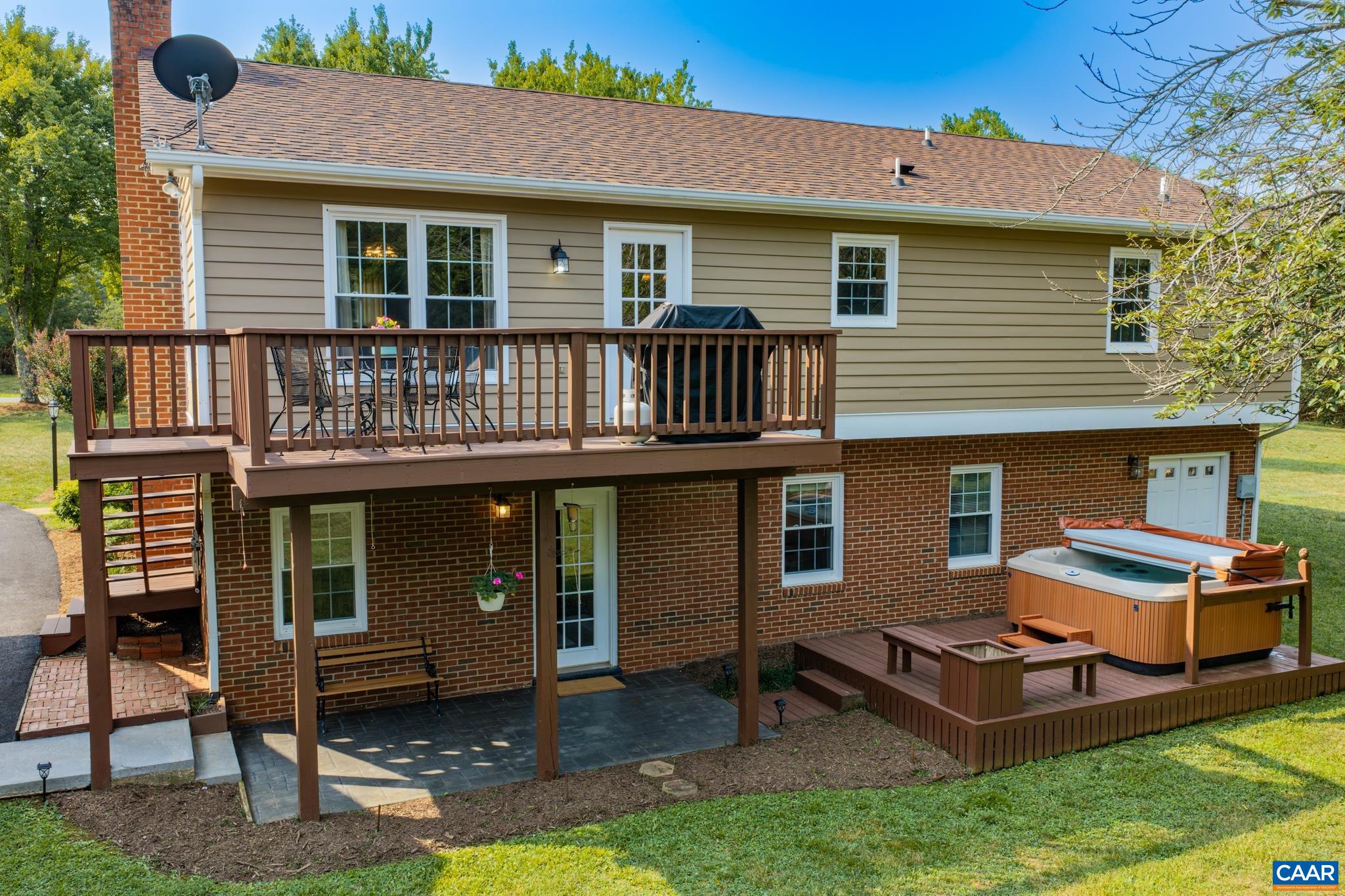 A backyard retreat designed for ultimate entertaining! Features a multi-level wooden deck with space for outdoor dining, a covered patio, classic brick accents, and a private hot tub area surrounded by lush greenery. Endless outdoor living awaits!