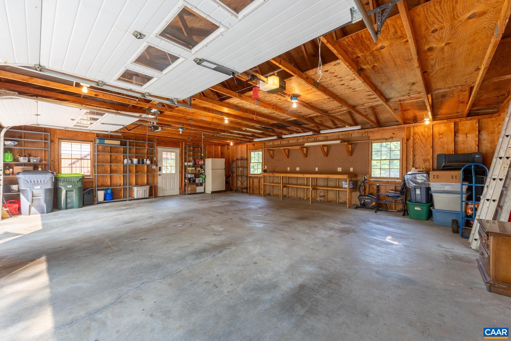 An incredible garage workshop setup! Features expansive concrete flooring, built-in wooden workbenches, extensive shelving, windows for natural light, and a personal entry door. Ultimate space for DIY projects, vehicle storage, and organization!