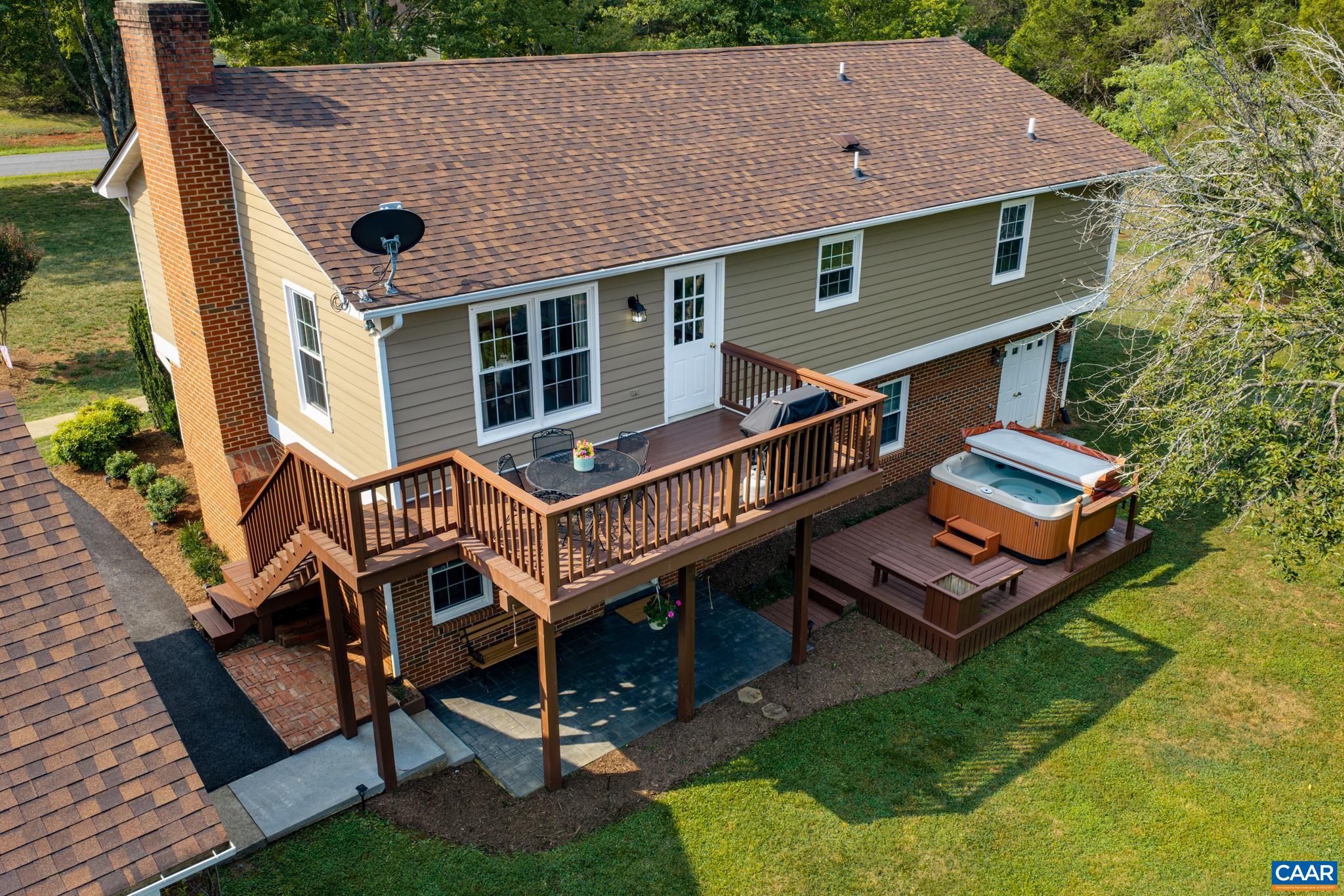 Stunning aerial view of an outdoor entertainer's paradise! Highlights include a multi-level wooden deck, private hot tub, covered patio below, classic brick chimney, and lush green surrounds. Ideal for seamless indoor-outdoor living!