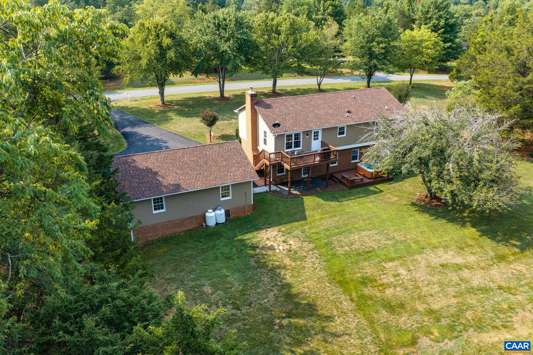 Stunning aerial view of this peaceful countryside property! Highlights include a multi-level deck with a hot tub, lower patio, attached garage, and a expansive green lawn framed by mature, lush trees. Your private outdoor sanctuary awaits!