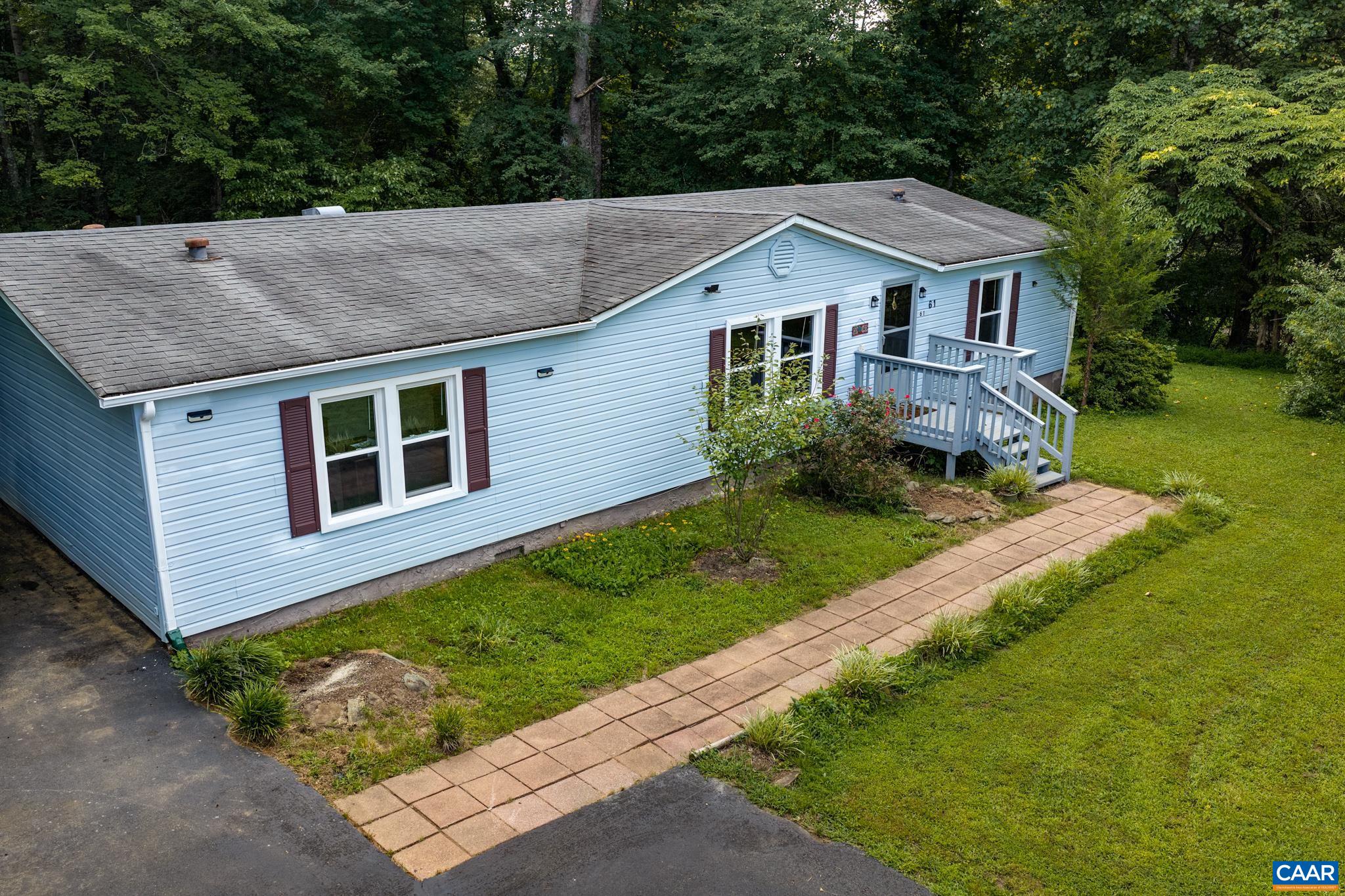 Elevated view highlighting serene light blue siding, crisp dark shutters, and a long paver walkway leading to the porch! Nestled on a green lawn against a lush, wooded backdrop, this home delivers charming curb appeal and quiet privacy.