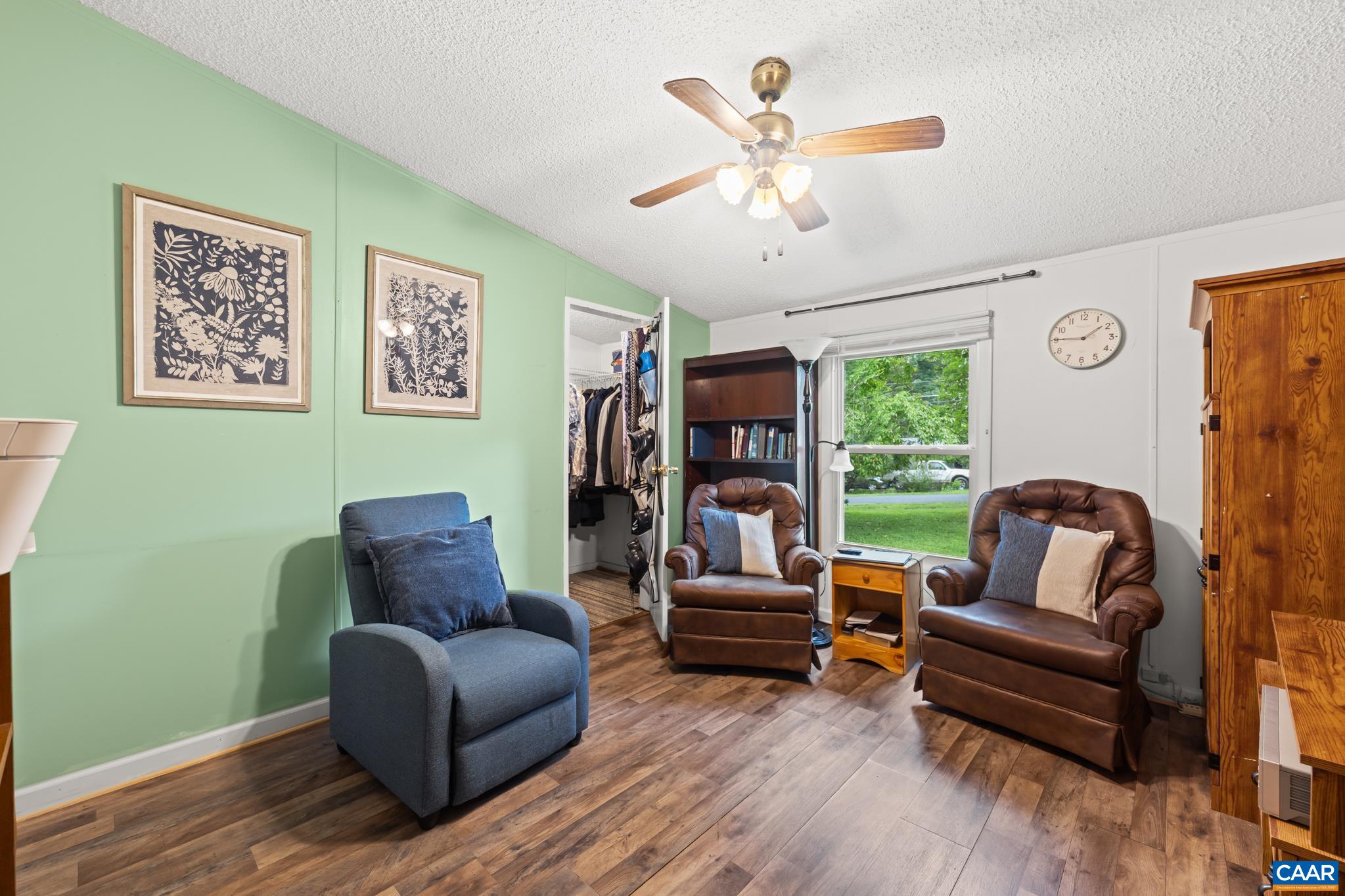 Cozy and versatile sitting room or bedroom featuring stylish sage green accent walls, rich wood-look flooring, and a ceiling fan. Bathed in natural light with peaceful views, it includes a walk-in closet for optimal storage and everyday comfort!