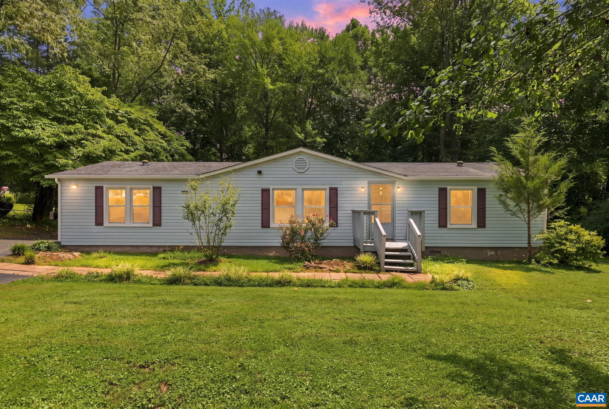 Lovely single-story home framed by mature trees and a vibrant lawn! Features warm exterior lighting, charming dark shutters, a covered entry porch, and dusk-lit windows that create an irresistible, move-in-ready welcome.