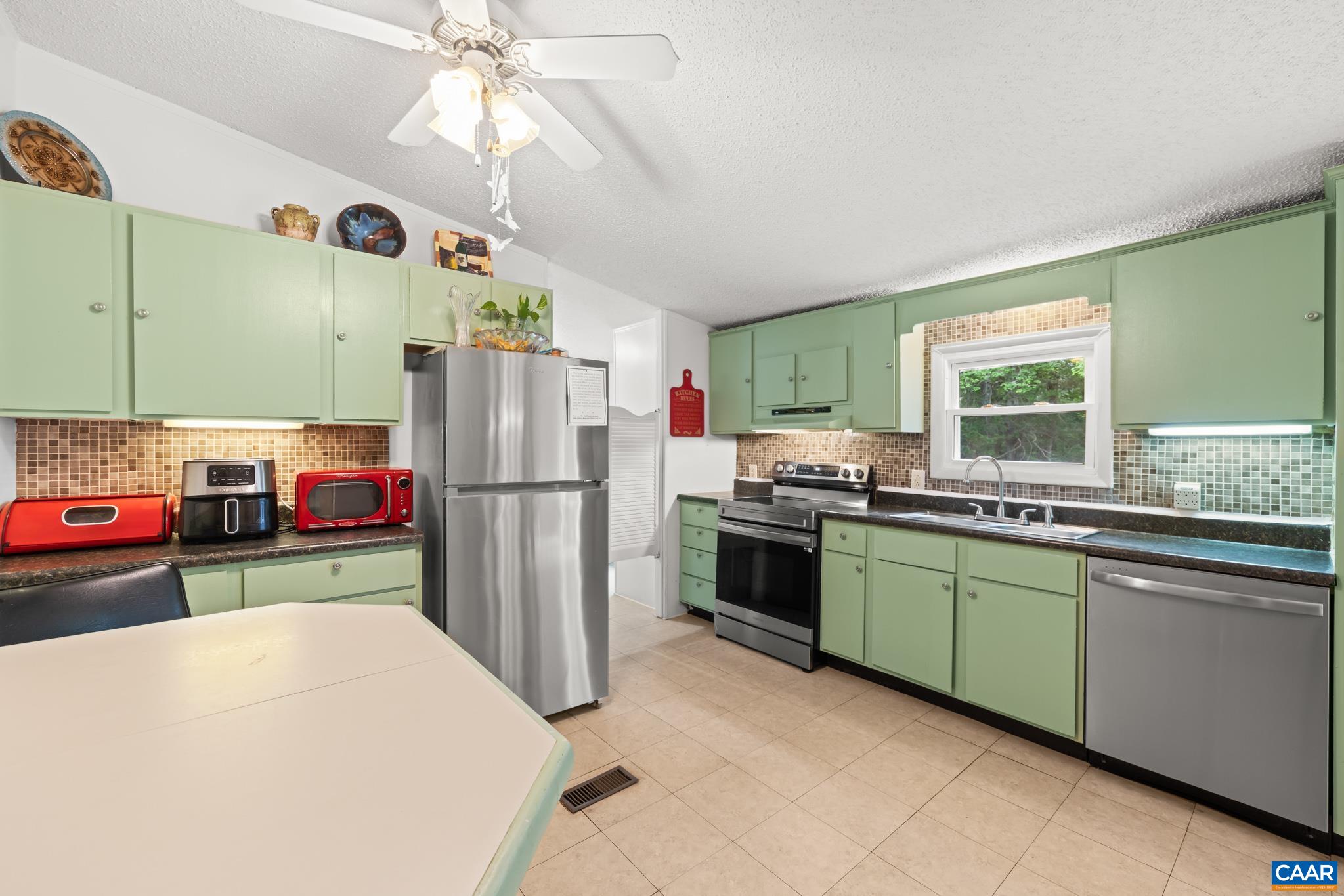 Charming and sunlit kitchen featuring vintage sage green cabinetry, updated stainless steel appliances, a stylish mosaic tile backsplash, and a functional island/peninsula with meal-prep space. A delightful heart of the home!