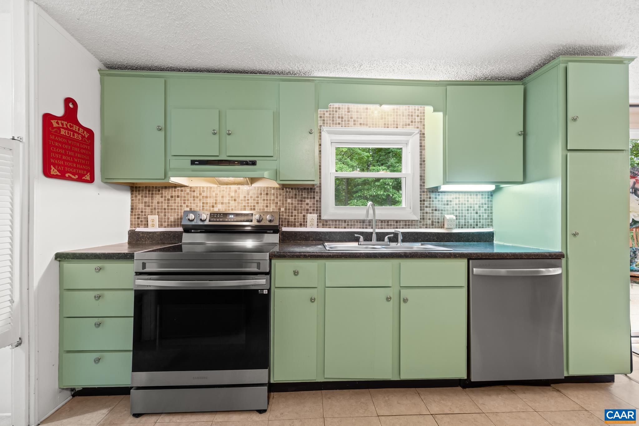 Bright and cheerful kitchen featuring charming sage green cabinetry, a stylish mosaic backsplash, and updated stainless steel appliances. Sunlit and functional, it offers ample storage and prep space for all your culinary creations!