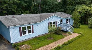 Elevated view highlighting serene light blue siding, crisp dark shutters, and a long paver walkway leading to the porch! Nestled on a green lawn against a lush, wooded backdrop, this home delivers charming curb appeal and quiet privacy.
